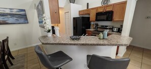 Eine Küche mit Granit-Arbeitsplatten, einem Kühlschrank, einer Mikrowelle und einem Herd. Zwei Stühle und eine kleine Kücheninsel mit Spüle sind ebenfalls vorhanden.