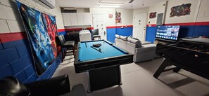 A room with a pool table, a sofa, a foosball table, and a TV displaying a streaming service. - Luxury Disney Vacation Home Private Pool Resort Amenities Sleeps 8 Game Room (Kissimmee)