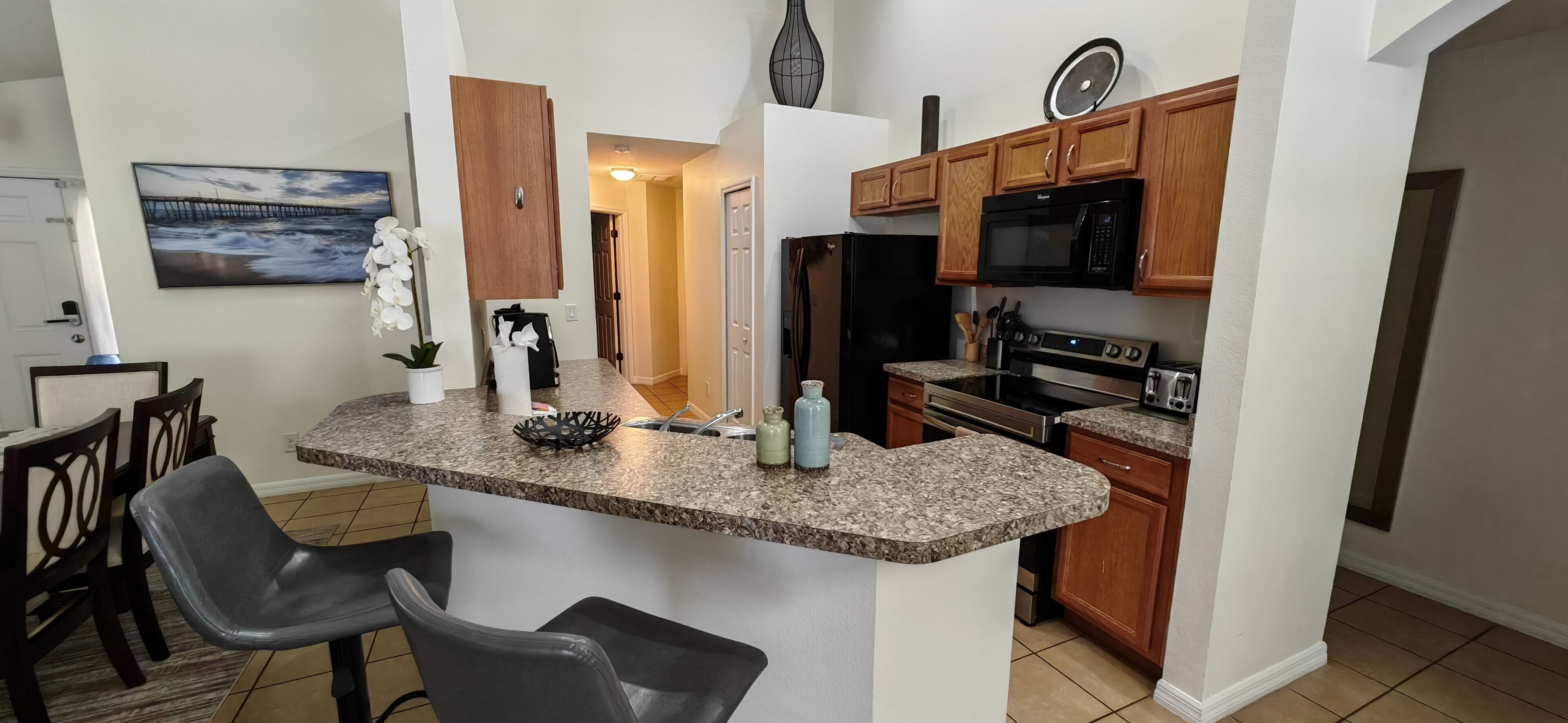 A compact kitchen with granite countertops, wooden cabinets, a microwave, stove, and a sink. There are two chairs and a potted orchid.