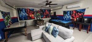 Ein Zimmer mit Pooltisch, einer Couch mit Kissen, einem Kicker und großen Wandbildern mit Superhelden-Motiven.