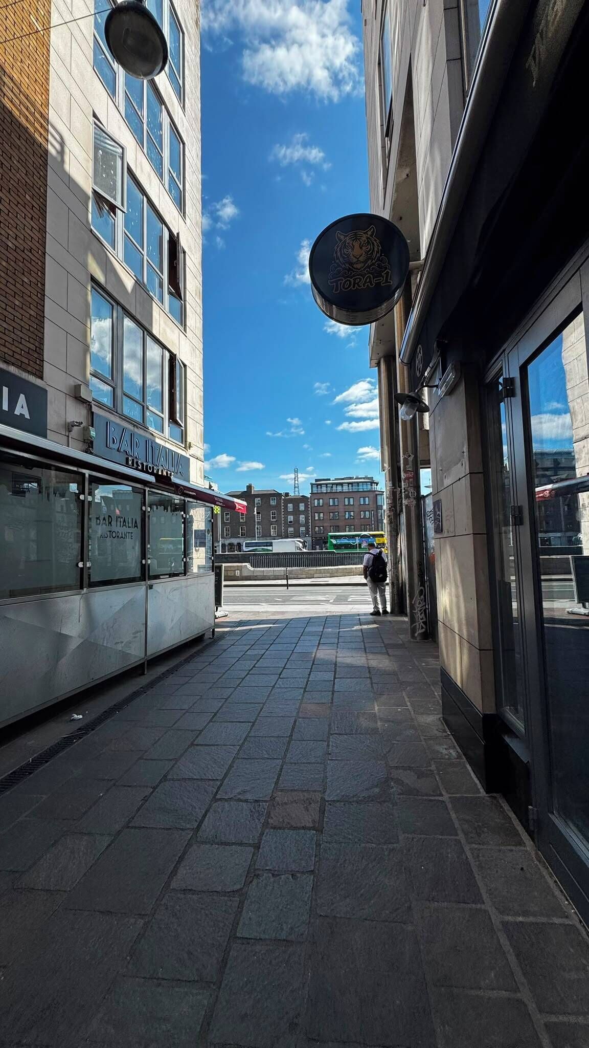 Een voetpad met een restaurant genaamd 'BAR ITALIA' aan de linkerkant en een gebouw met een bord 'TORA-1' aan de rechterkant.