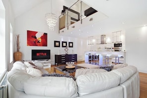 A modern living room with a large sofa, a TV, and a dining area with bar stools. - Luxury 3-Bed Penthouse · Sleeps 7 · Gated · NEC (Shirley)