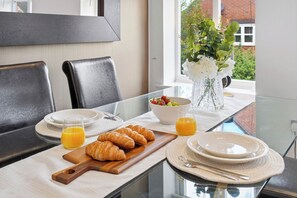 A breakfast setting with croissants, fruit, and orange juice on a glass table with a view of greenery outside. - Luxury 3-Bed Penthouse · Sleeps 7 · Gated · NEC (Shirley)