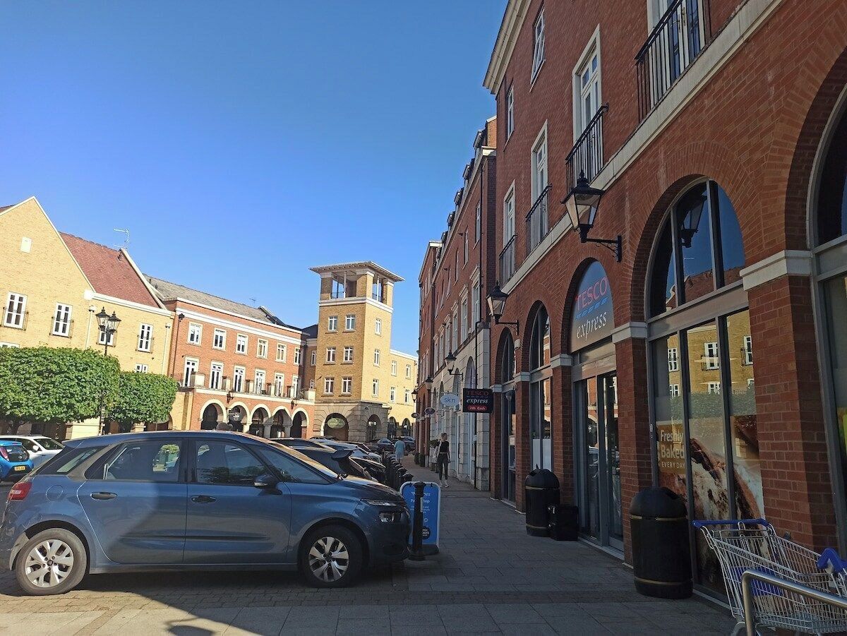 Une rue bordée de voitures garées, un magasin Tesco Express et un immeuble doté d’une tour.