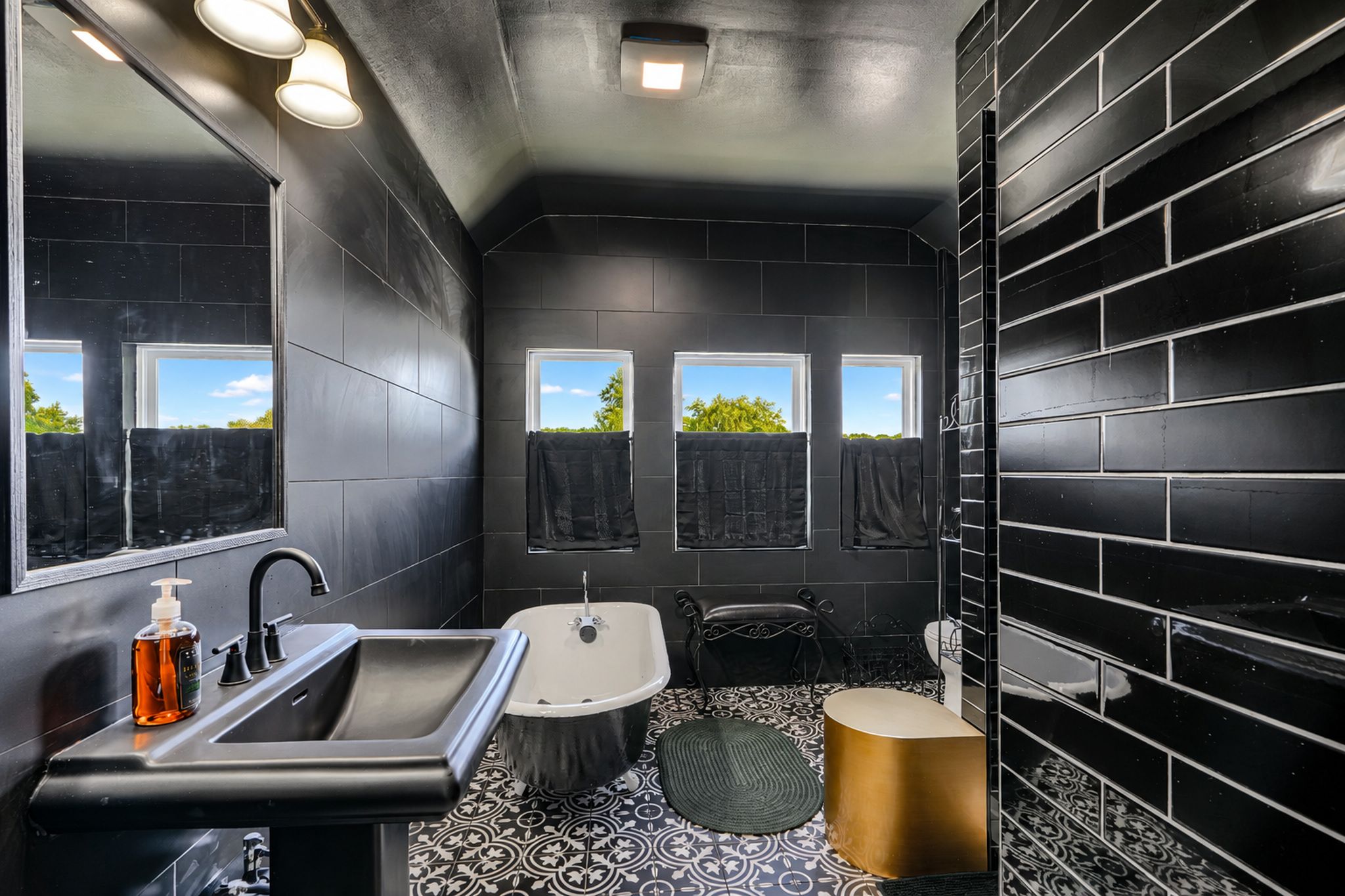 A modern bathroom with a black and white tiled floor, a sink, a bathtub, and a toilet. There are windows allowing natural light and views of greenery outside.