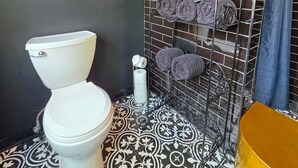 Ein Badezimmer mit einer weißen Toilette, einer Wand mit schwarzen Fliesen und einem Regal mit aufgerollten Handtüchern.