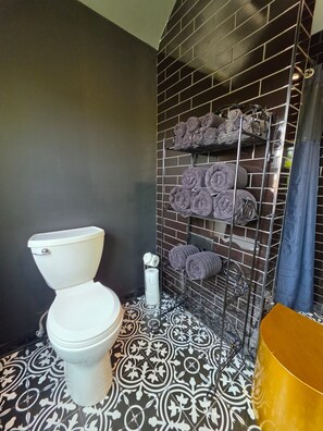Une salle de bain avec des toilettes blanches, un mur recouvert de carreaux noirs et une étagère où sont pliés des serviettes.
