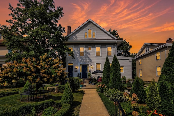 A two-story house with a front porch, a pathway leading to the entrance, and a garden with various plants and a bench.