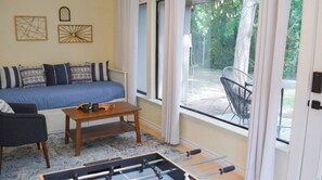 Ein Zimmer mit einer Couch, einem Couchtisch und einem Kicker. Zwei Fenster mit Vorhängen und Blick ins Freie.