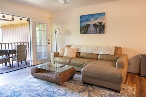 A living room with a glass coffee table, a beige sofa, and a painting of a beach scene. - Beautiful Beach Bungalow (Kihei)
