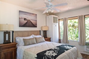 A bedroom with a bed, a ceiling fan, a nightstand with a lamp, a picture on the wall, and a window with curtains. - Beautiful Beach Bungalow (Kihei)