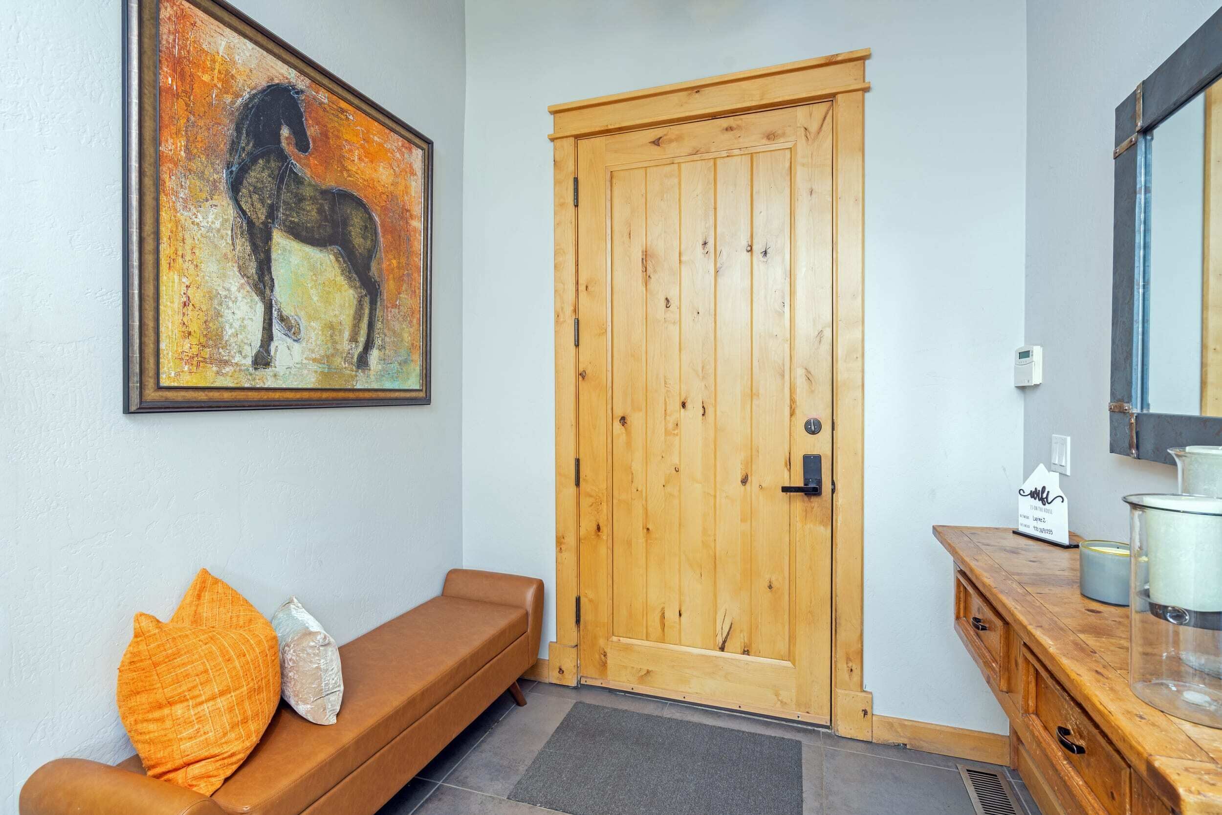 A room with a wooden door, a bench with cushions, a painting of a horse, and a mirror on the wall.