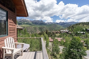 Eine Holzhütte mit einer Terrasse und einem Stuhl, mit Blick auf ein Tal, in dem Berge und eine kleine Siedlung liegen.