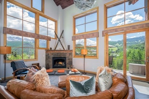 Ein geräumiges Wohnzimmer mit Ledersofas, einem Kamin und großen Fenstern mit Blick auf die Berge.