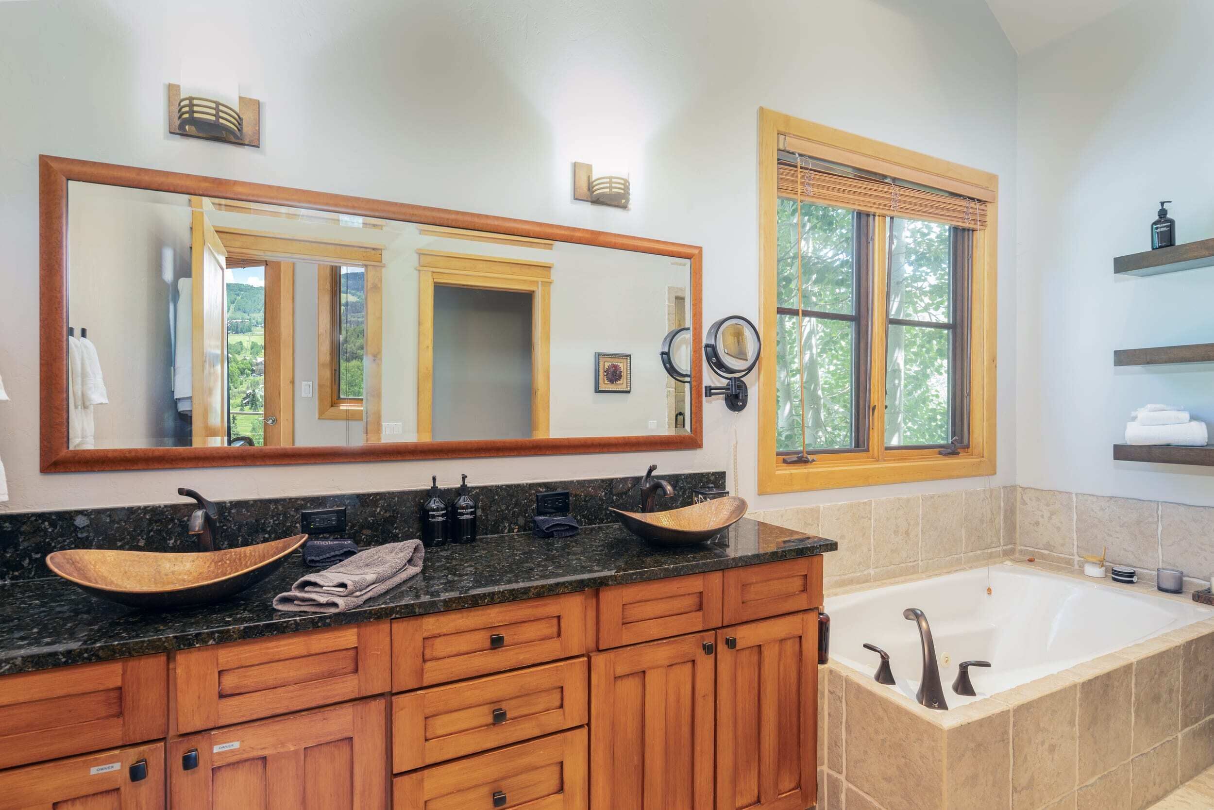 A bathroom with a large mirror, wooden cabinets, a vessel sink, a bathtub, and a window with blinds.