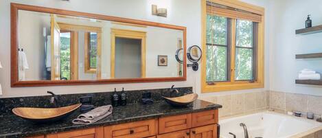 A bathroom with a large mirror, wooden cabinets, a vessel sink, a bathtub, and a window with blinds.
