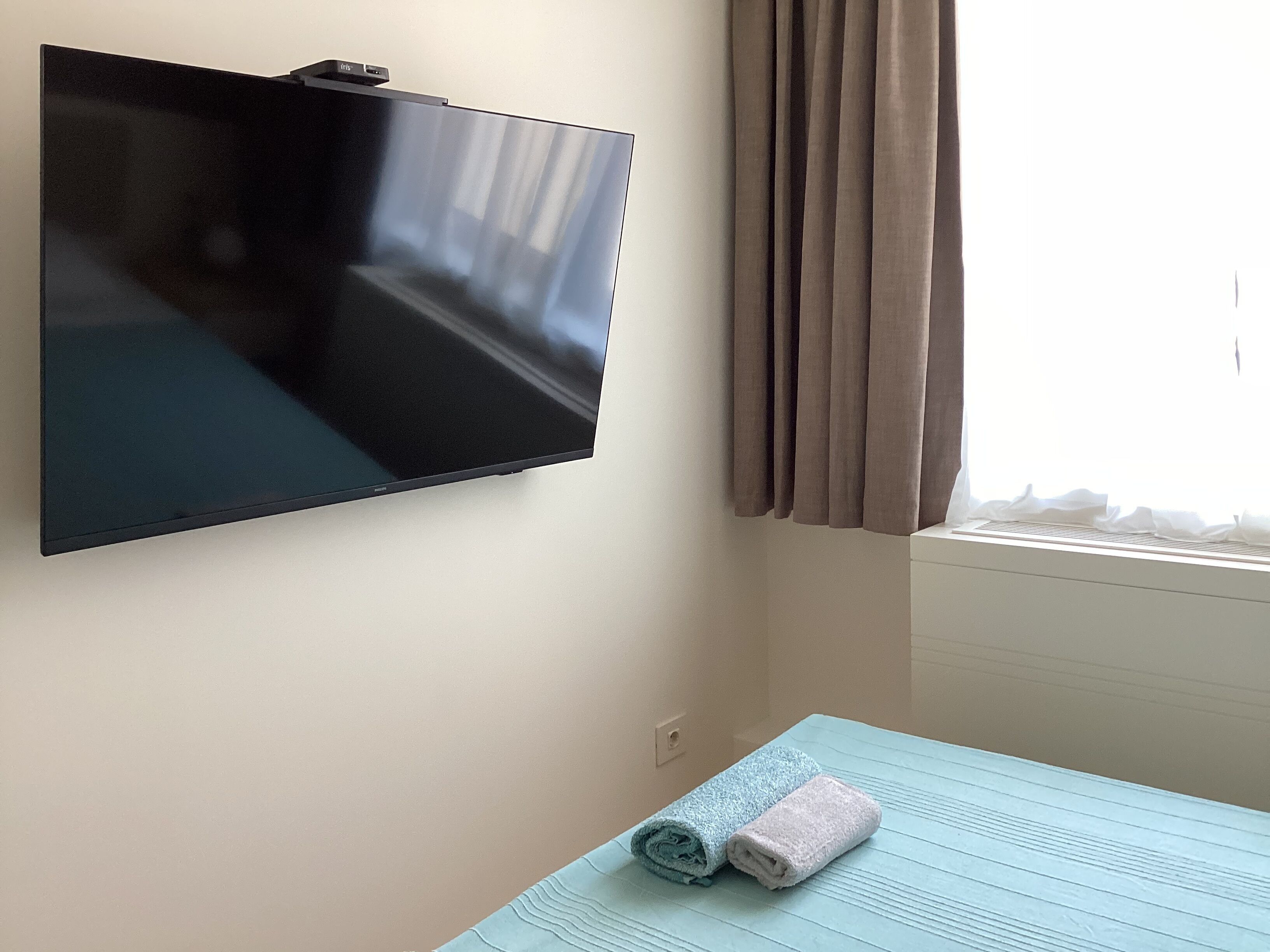 A room with a flat-screen TV mounted on the wall, a window with curtains, and a bed with two rolled towels.