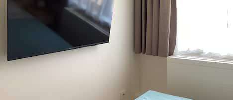 A room with a flat-screen TV mounted on the wall, a window with curtains, and a bed with two rolled towels.