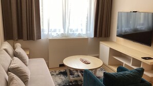 Una habitación de hotel moderna con un sofá, una mesita redonda pequeña, un televisor y una ventana con cortinas.