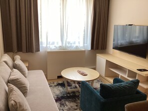 Et moderne hotelværelse med sofa, rundt bord, fjernsyn og et vindue med gardiner.