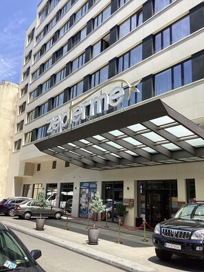 Entrada de hotel con fachada moderna, zona de acceso cubierta y estacionamiento con coches y plantas en macetas.