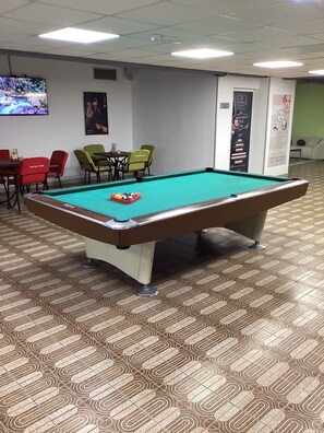 A pool table with a green felt surface, surrounded by patterned carpet flooring, and a television mounted on the wall.