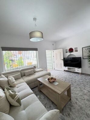 A modern living room with a sectional sofa, a wooden coffee table, a television, and a large window with a view of greenery. - Townhouse in a quiet neighborhood (El Chaparral)