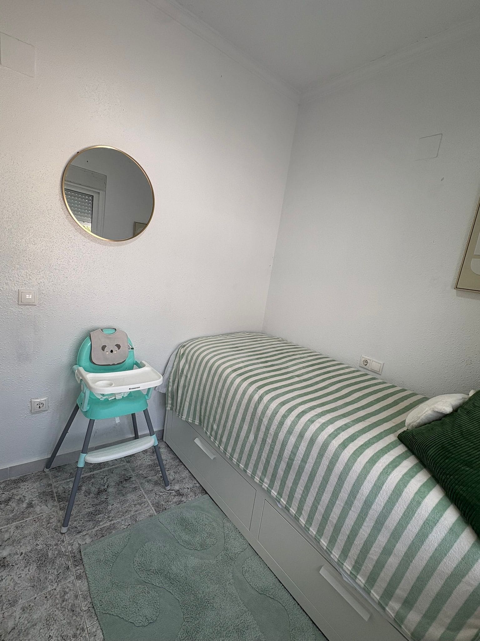 Une petite chambre avec un lit simple, une couverture à rayures, un miroir rond accroché au mur et une chaise haute appuyée contre le mur.