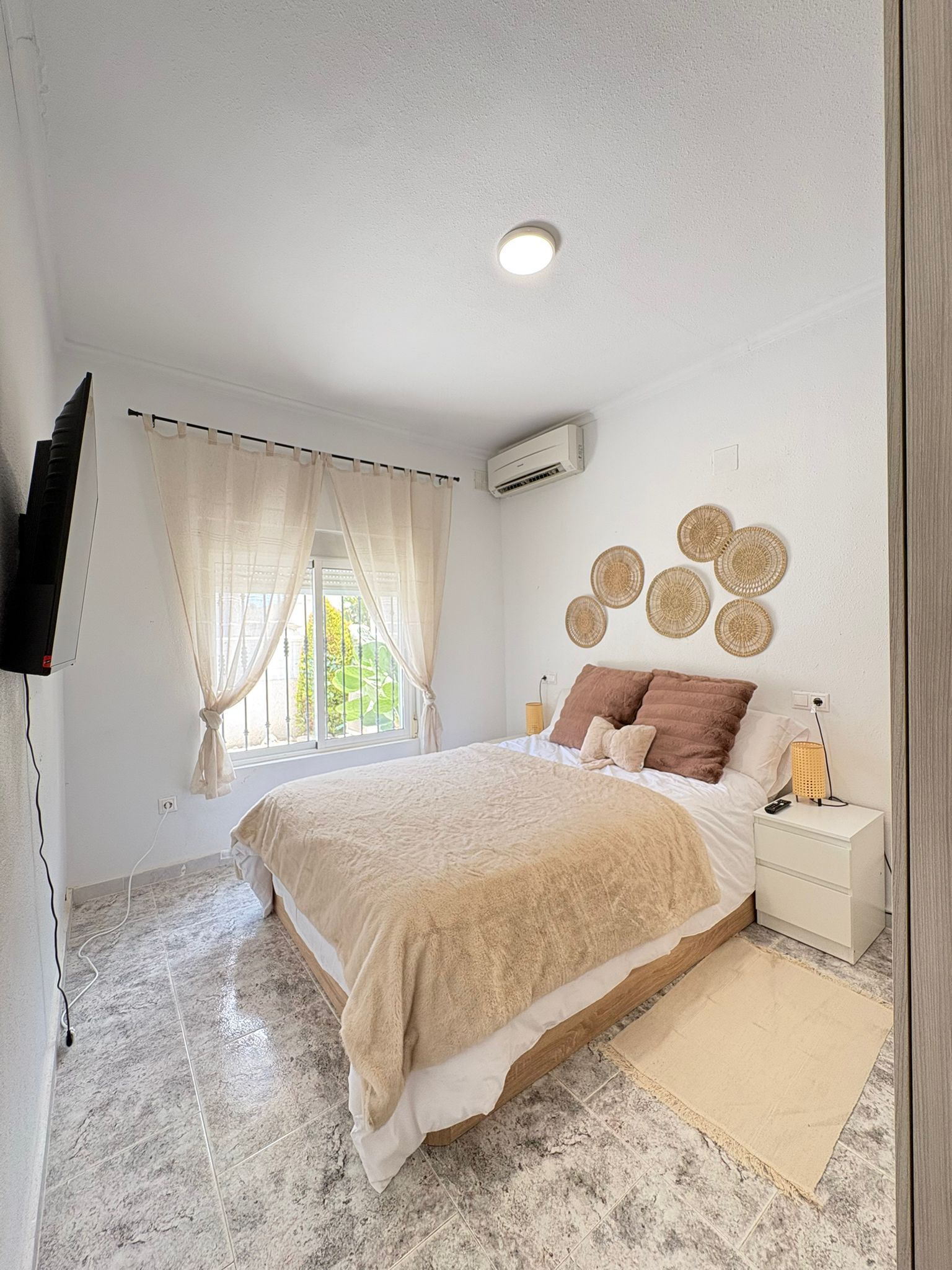 A bedroom with a bed, a nightstand, a wall-mounted air conditioner, and a window with curtains.