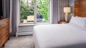 Hotellihuone, jossa on sänky, puinen lipasto, yöpöytä lampun kanssa ja ikkuna, jossa on verhot.