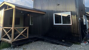A two-story building with a corrugated metal exterior and a wooden deck. - Casita Nalca / Canasto de Agua Huts—Chaitén, Chilean Patagonia. (Chaiten)
