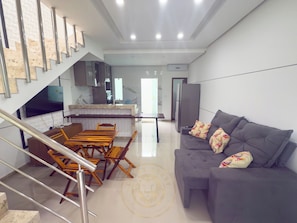 A modern living room with a grey sofa, wooden dining table, and chairs. A staircase with metal railings leads to an upper level. - Taperapuã Beach - Ashby Houses (Porto Seguro)