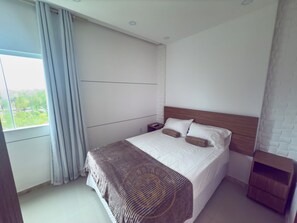 A hotel room with a large bed, a nightstand, a wooden headboard, and a view of greenery outside. - Taperapuã Beach - Ashby Houses (Porto Seguro)