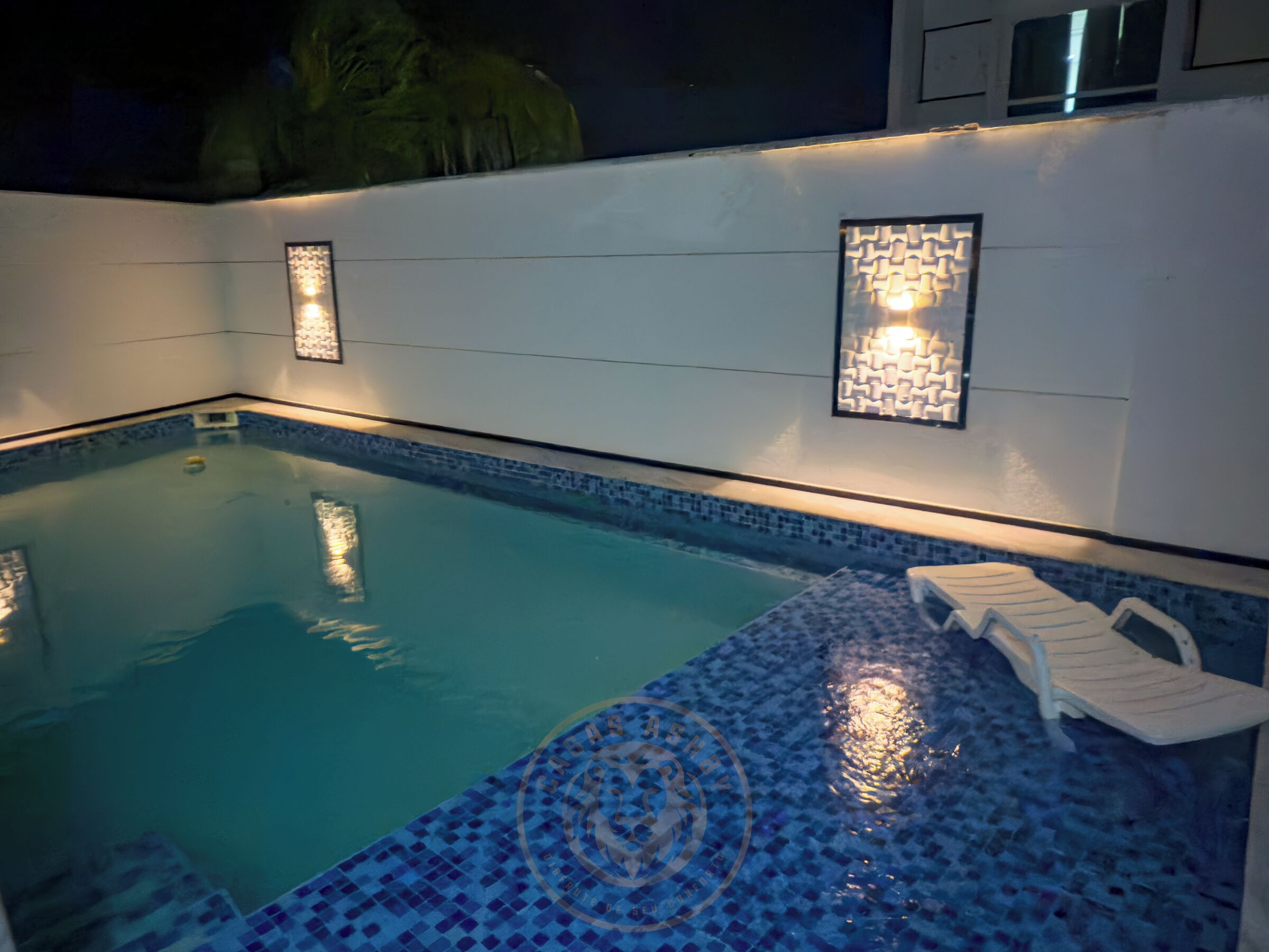Una piscina con borda piastrellata, una piccola scala e due luci a parete illuminate.