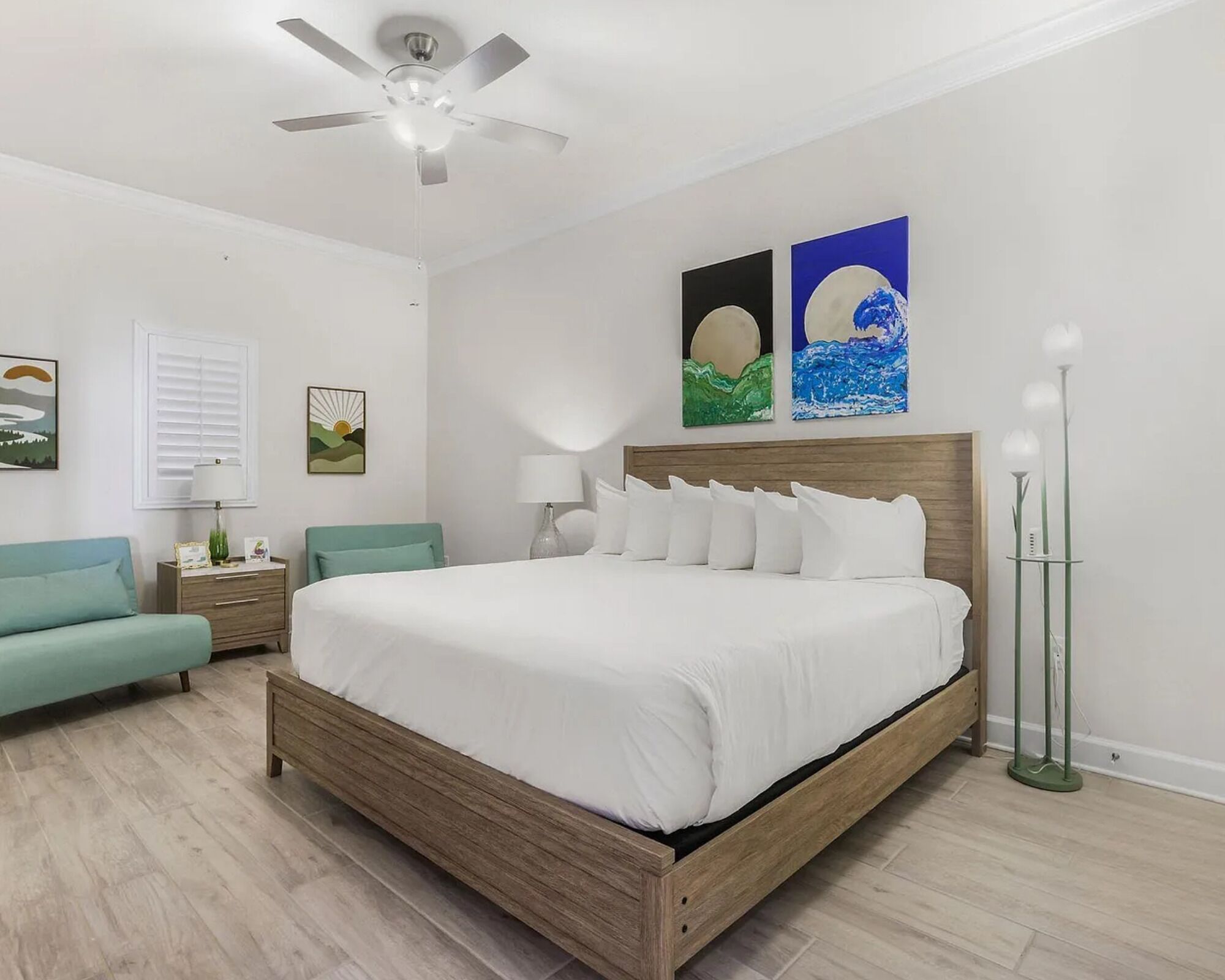 A bedroom with a wooden bed, a ceiling fan, a nightstand, a lamp, a bedside table, a green chair, and artwork on the wall.