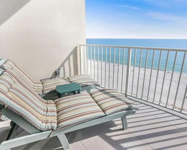A balcony with a view of the beach, a seating area with striped cushions, and a small table. - Phoenix GS II #1003 - Luxury Beachfront 3BR, Lazy River, Heated Pool, Hot Tub (Gulf Shores)