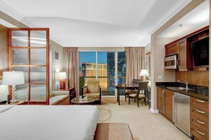 A hotel room with a bed, a sitting area with a table and chairs, a kitchenette, and a large window offering a city view. - MGM Signature-27-705 F1 Track & Strip View Balcony (Las Vegas)
