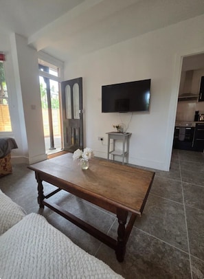 A living room with a wooden dining table, a flat-screen TV mounted on the wall, and a kitchen in the background.