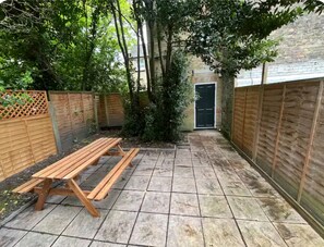 A wooden bench in a garden area with a stone-tiled floor, surrounded by wooden fences and a brick wall.