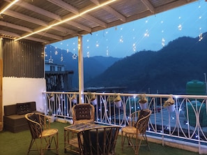 A balcony with wicker furniture, string lights, and a view of mountains. - The homez haven (Rishikesh)