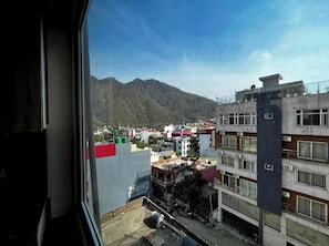 A view from a window overlooking a cityscape with mountains in the background, featuring multi-story buildings with various architectural styles. - The homez haven (Rishikesh)