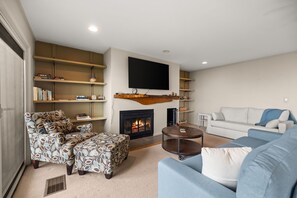 A living room with a fireplace, a flat-screen TV mounted above it, a sofa, a patterned armchair, and a coffee table. - Loggers Lookout Condo at The Homestead (Glen Arbor)