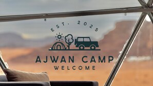 Une vue à travers la fenêtre d’une pièce où l’on aperçoit un lit, des oreillers et une pancarte sur laquelle on peut lire « AJWAN CAMP » et « BIENVENUE ».