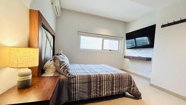 Ein Schlafzimmer mit Bett, Nachttisch, Lampe, Fenster und Wand-Fernseher.