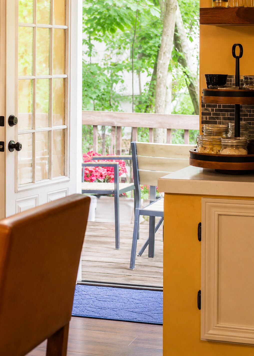 Um deck de madeira com um banco e uma mesa, uma porta que dá para um jardim, e uma área de cozinha com bancada e armários.