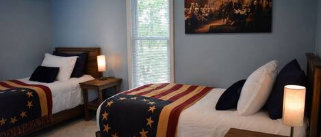A bedroom with two beds, each with a star-patterned blanket, a painting on the wall, and a window with blinds.