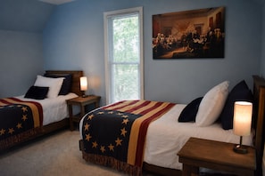 A bedroom with two beds, each with a star-patterned blanket, a painting on the wall, and a window with blinds.