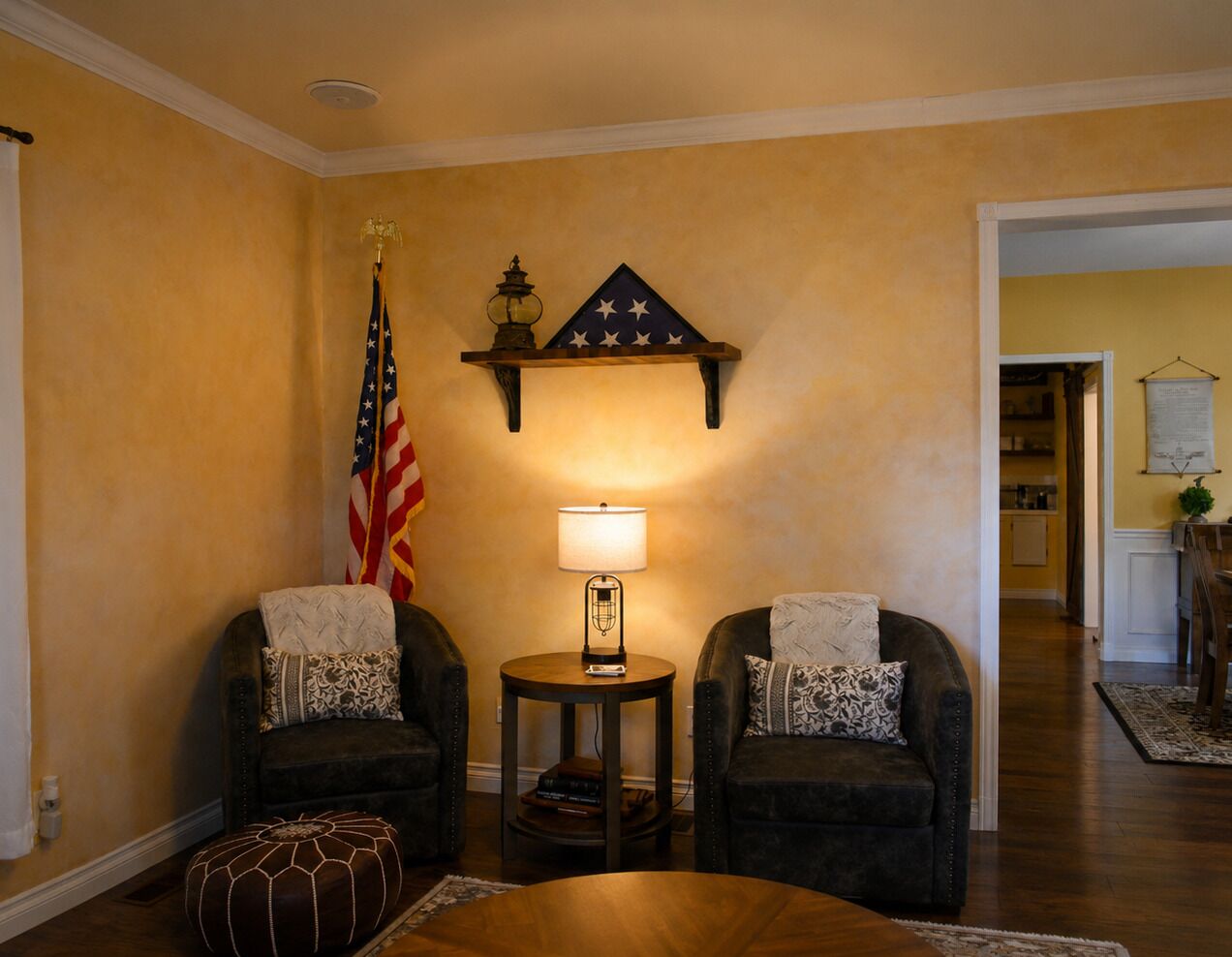 Uma sala de estar com duas poltronas, uma mesa de centro redonda, um abajur, uma bandeira e uma prateleira na parede com um objeto decorativo.
