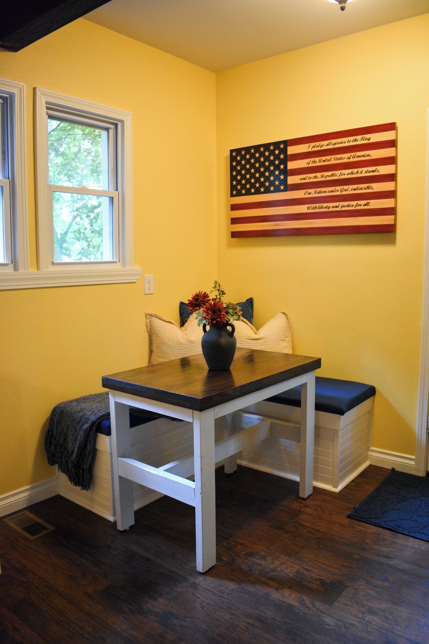 A room with a wooden table, a vase with flowers, a window, and an American flag on the wall.