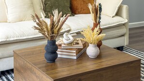 Uma sala de estar com sofá, almofadas decorativas, mesa de centro com livros e vasos, e obras de arte nas paredes.